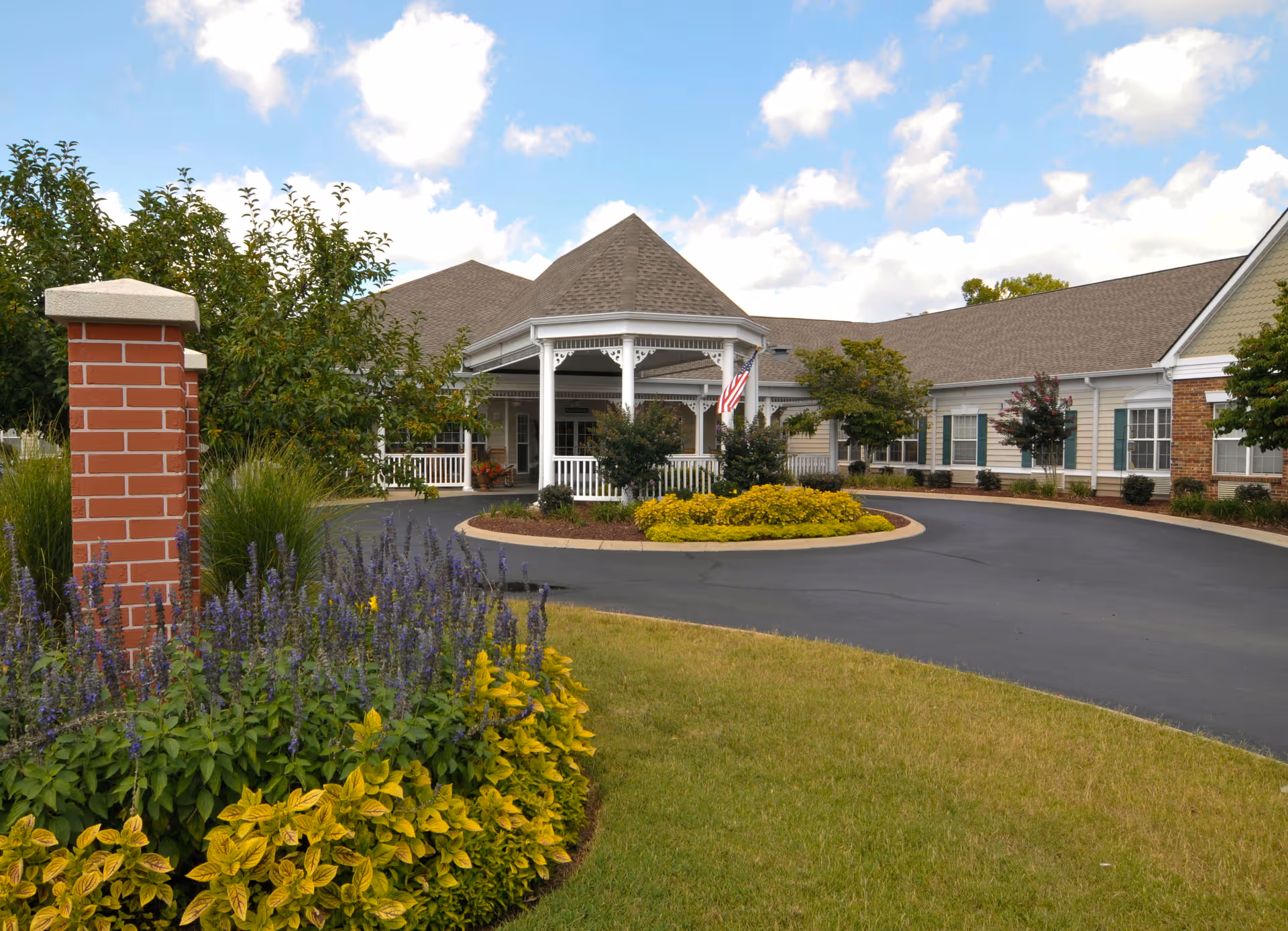 Exterior view of Charter Senior Living of Franklin showing a circular driveway with landscaped flower beds and shrubs. The building has a covered entrance with white columns and an American flag displayed. The sky is partly cloudy.