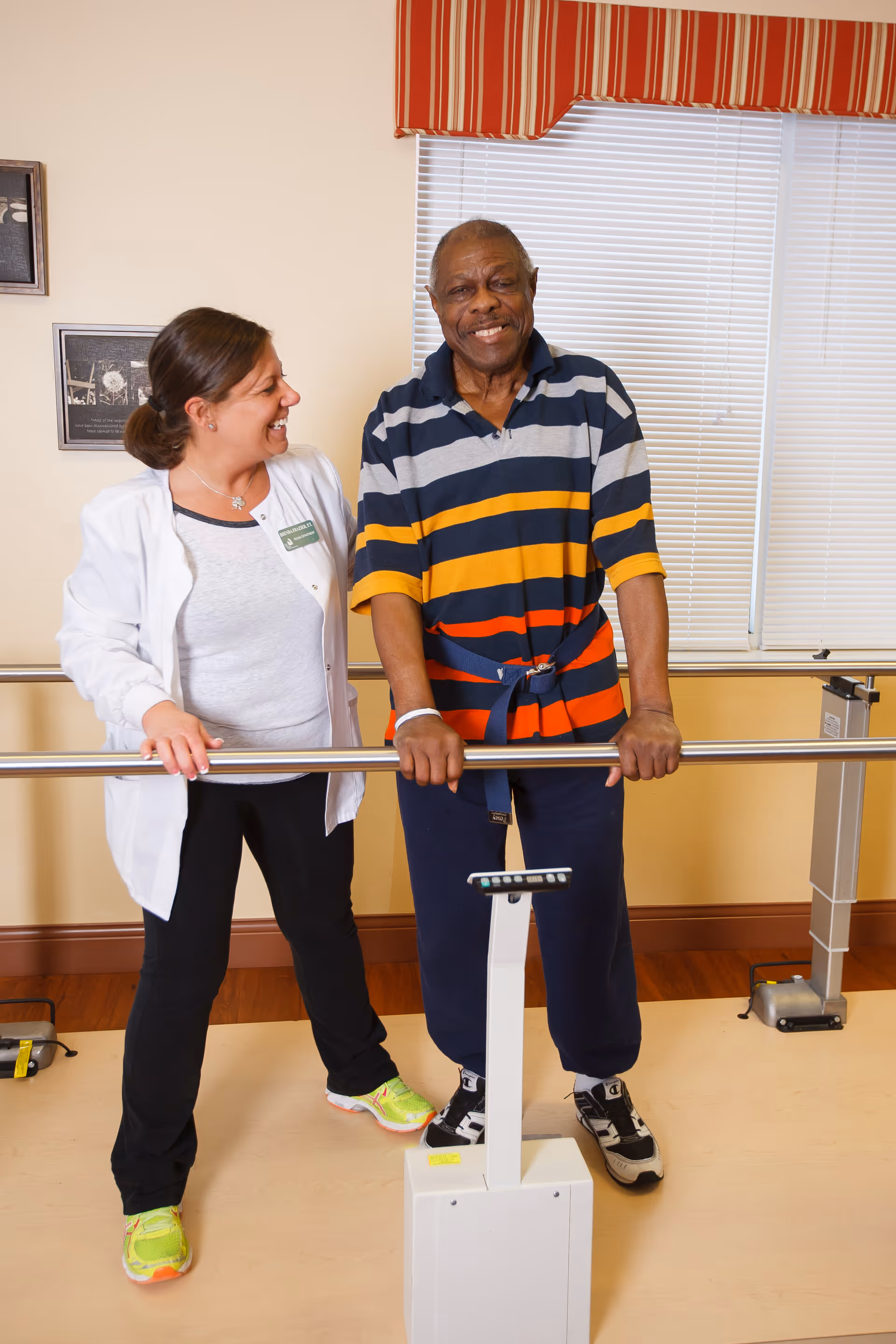 A senior man wearing a striped shirt and dark pants is standing and holding onto parallel bars for support during a physical therapy session. A female healthcare professional in a white coat and bright sneakers stands beside him, smiling and providing encouragement. The room has a window with blinds and a red-striped valance.