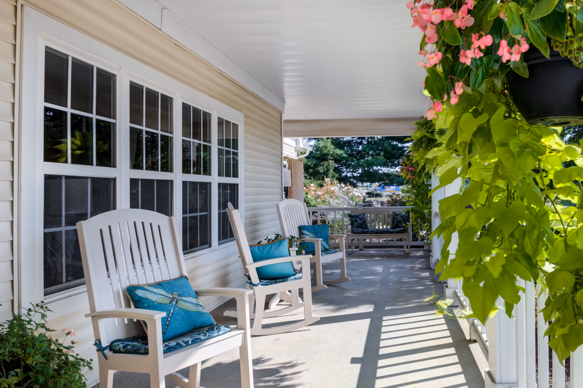 A covered porch area with white rocking chairs and a bench, each adorned with blue cushions. The porch is decorated with hanging plants and flowers, and sunlight casts shadows on the concrete floor. The porch is attached to a building with beige siding and large windows.
