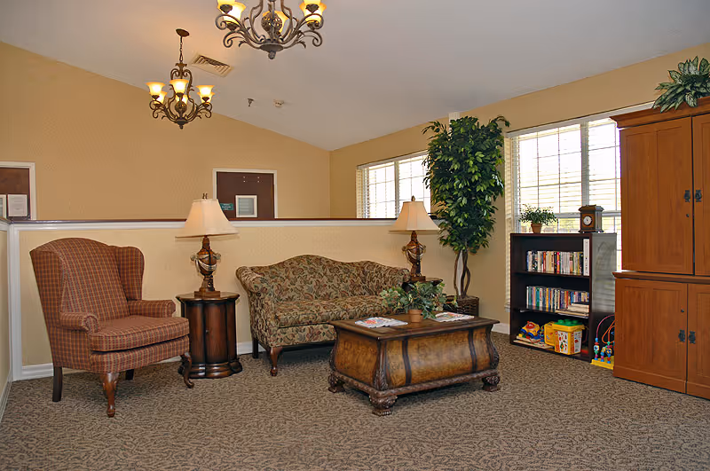 A cozy sitting area with a patterned armchair, a floral upholstered sofa, a wooden coffee table with magazines and a plant, two table lamps on side tables, a tall green plant near two windows, a bookshelf with books and toys, and a wooden cabinet. The room has beige walls, carpeted floor, and two decorative ceiling lights.