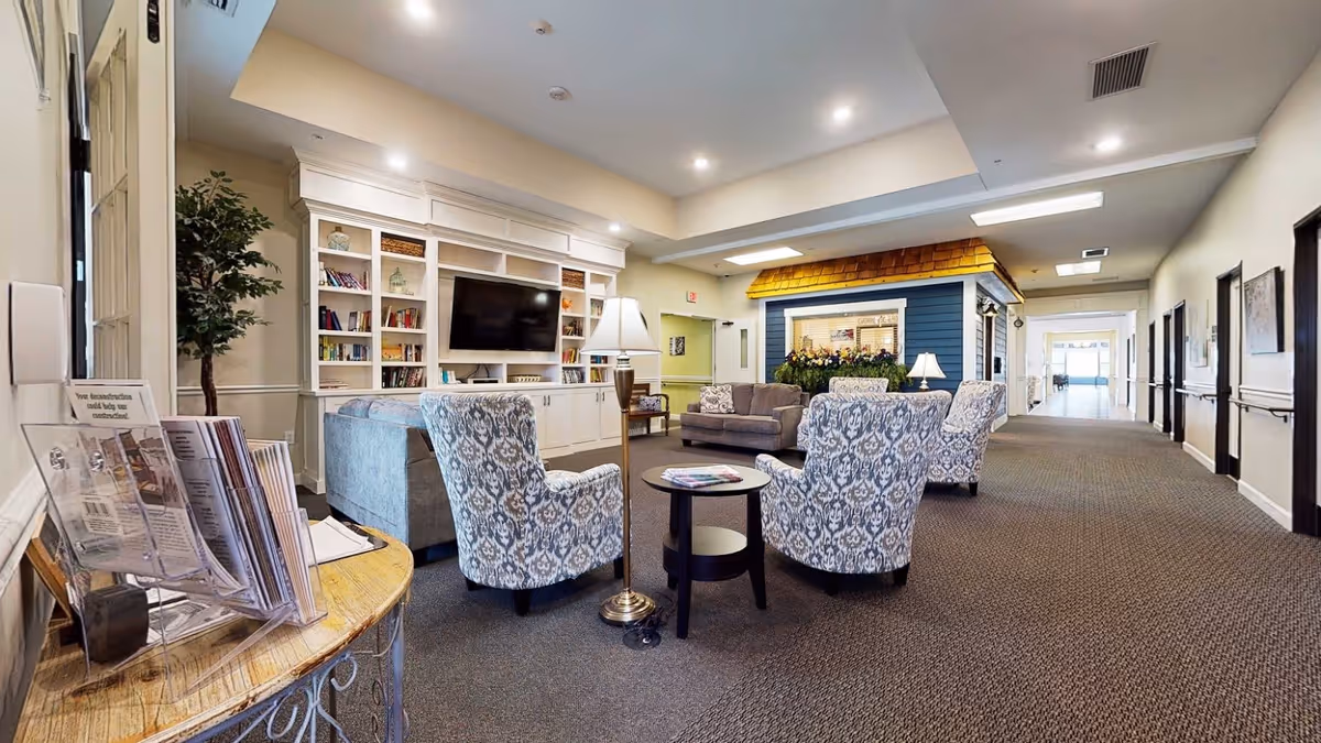 A spacious and well-lit common area in a senior living facility featuring patterned armchairs and sofas arranged around small tables. There is a large built-in white shelving unit with a flat-screen TV and decorative items. A small blue structure resembling a house with a shingled roof is visible in the background. The area has carpeted floors, lamps, and a hallway leading to other rooms.