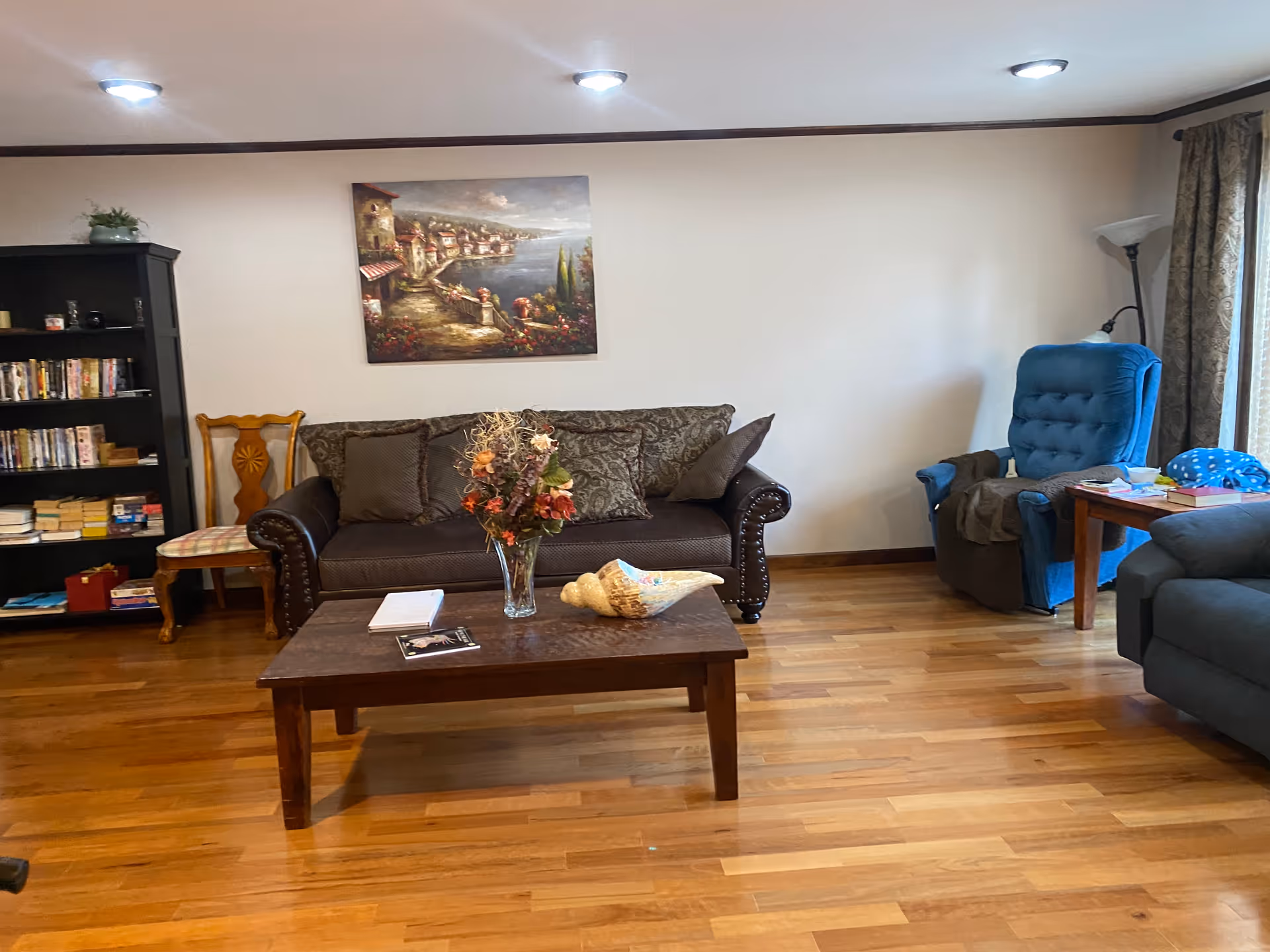 Living room with brown sofa, coffee table, bookshelf, blue recliner, and wall painting.