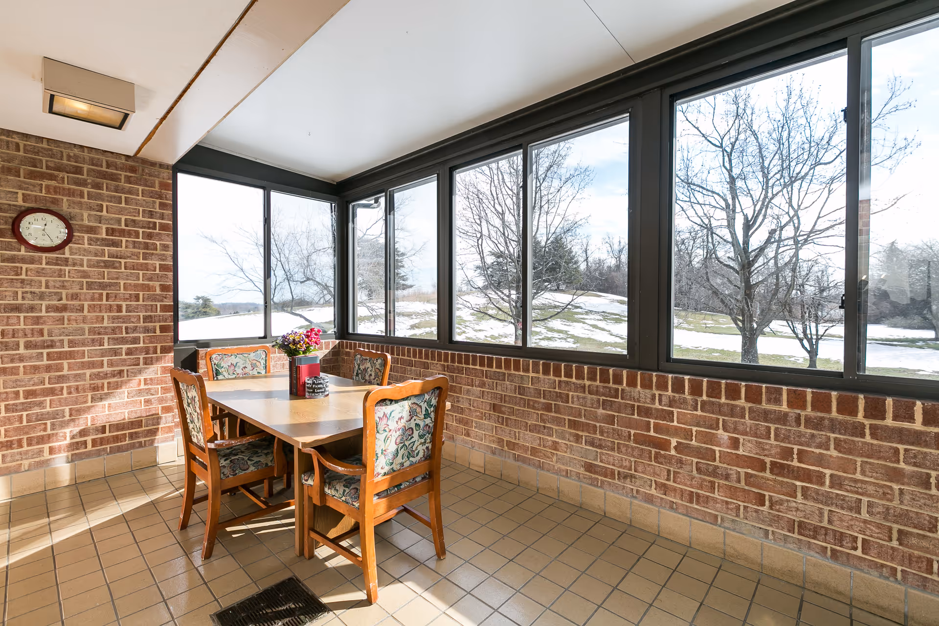 A sunlit enclosed patio area with large windows showing a snowy landscape outside. Inside, there is a wooden table with four floral upholstered chairs around it. A small flower arrangement and a red container are placed on the table. The walls are made of brick and the floor is tiled. A clock is mounted on the brick wall.