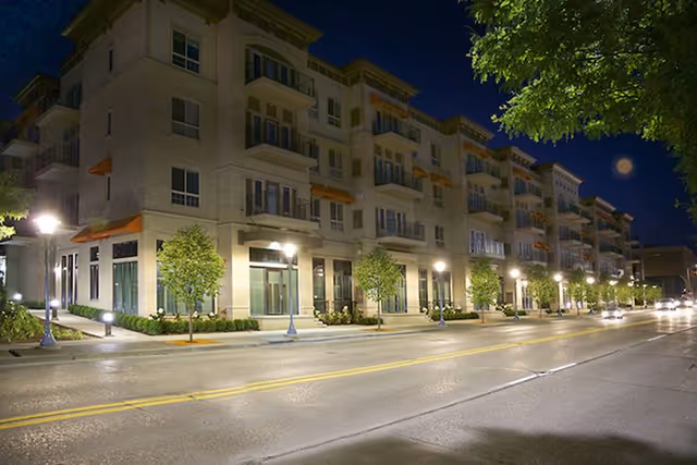 A multi-story residential building at night with street lamps illuminating the sidewalk and small trees lining the street. The building has balconies and large windows, with a clear sky and a visible moon in the background.