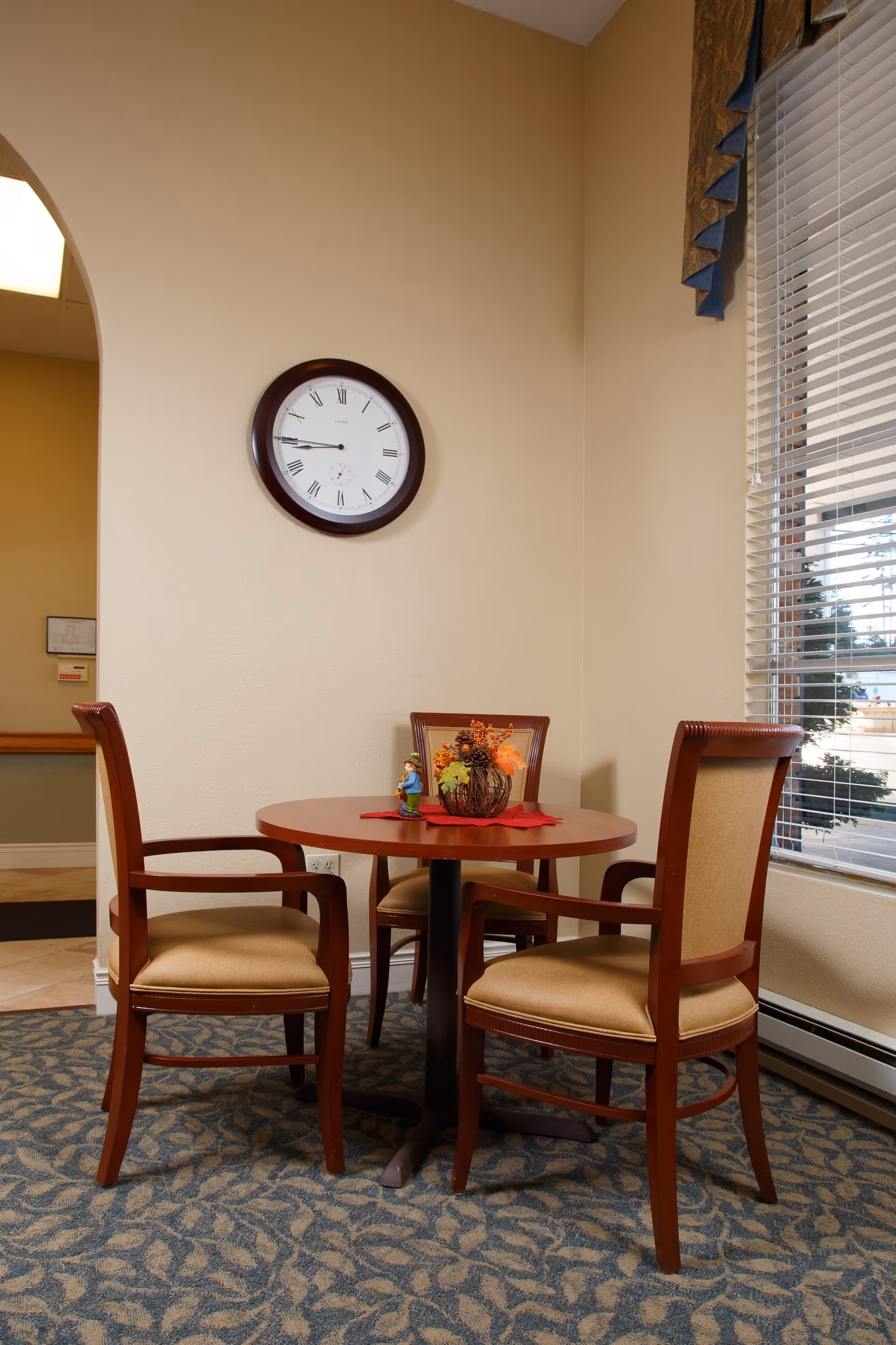 A small round wooden table with a decorative centerpiece and two wooden chairs with beige cushions in a room with beige walls. A round wall clock with Roman numerals hangs on the wall above the table. A window with blinds is on the right side, letting in natural light.