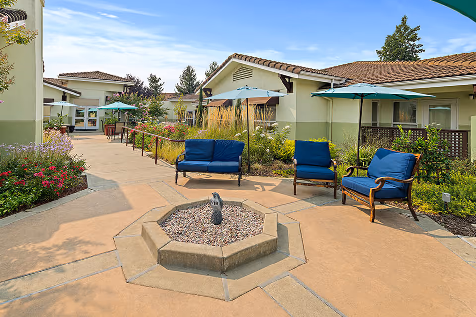 Outdoor patio area at The Meadows of Napa Valley with cushioned seating including a loveseat and chairs under blue umbrellas, surrounded by landscaped plants and flowers, with single-story buildings in the background under a clear sky.