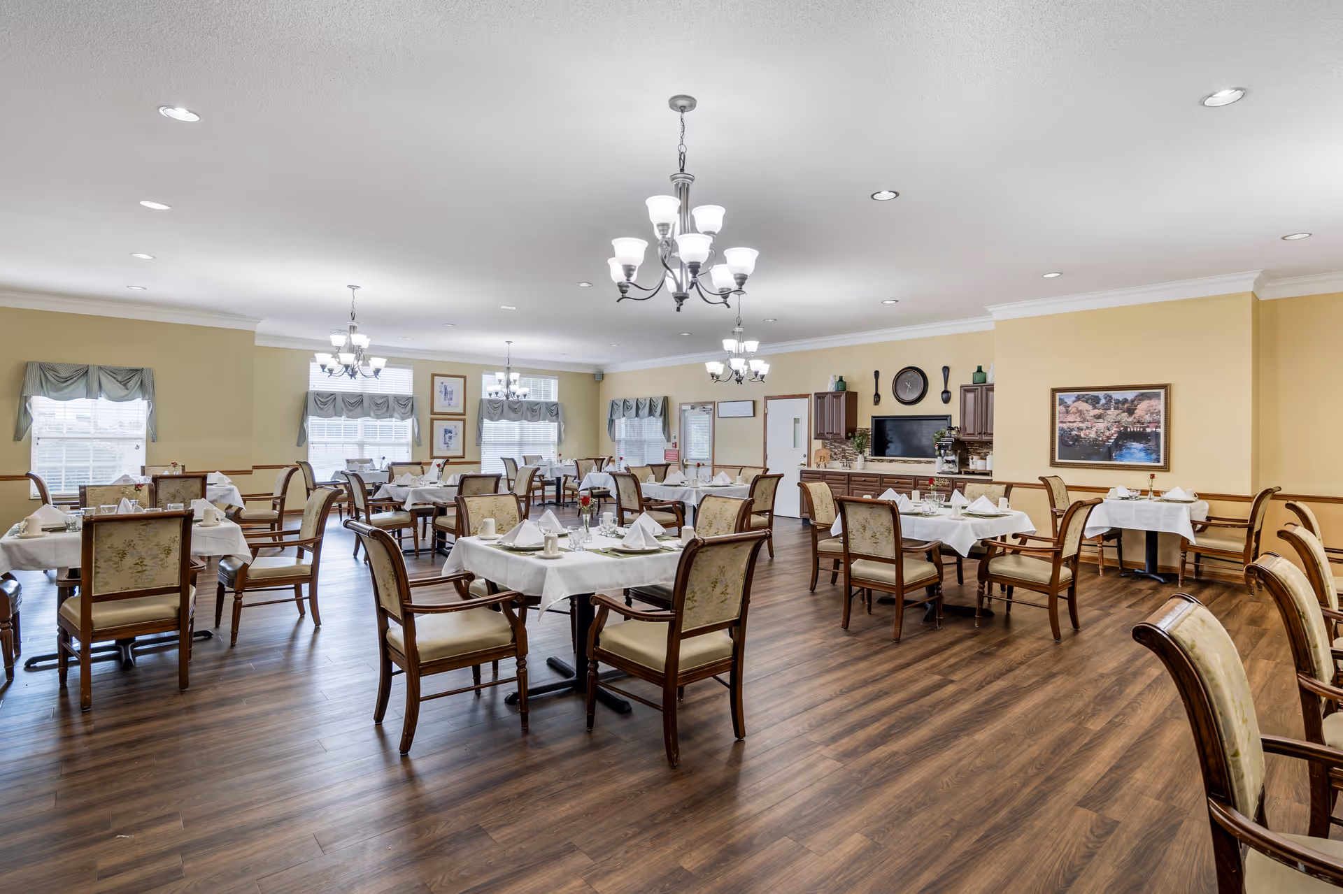 A spacious dining room with multiple tables covered in white tablecloths, each set with napkins, cups, and small flower vases. The room has wooden flooring, beige walls, several windows with valance curtains, and multiple chandeliers hanging from the ceiling. There is a cabinet with a TV and decorative items on the far wall.