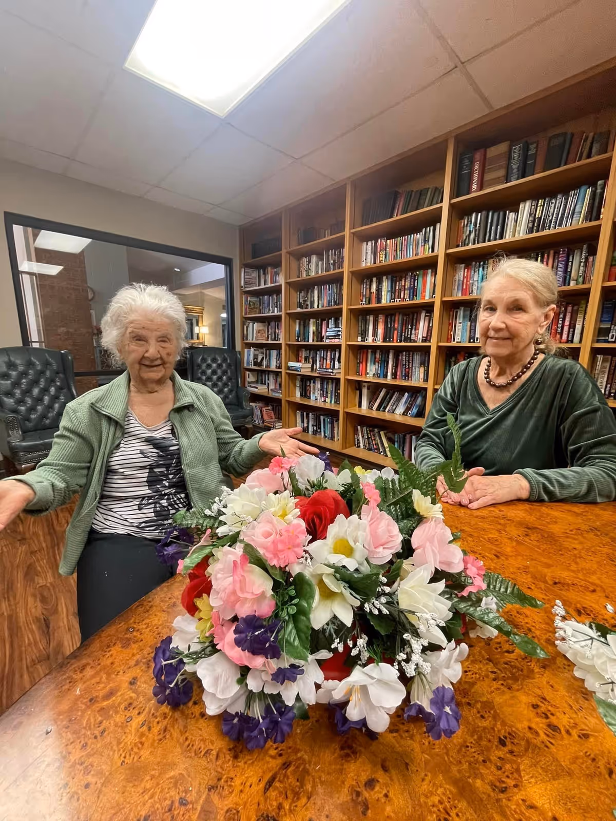 Two elderly women sitting at a wooden table with a large colorful bouquet of artificial flowers in front of them. Behind them is a wall filled with bookshelves containing many books. The room has a ceiling light and a window showing another room.