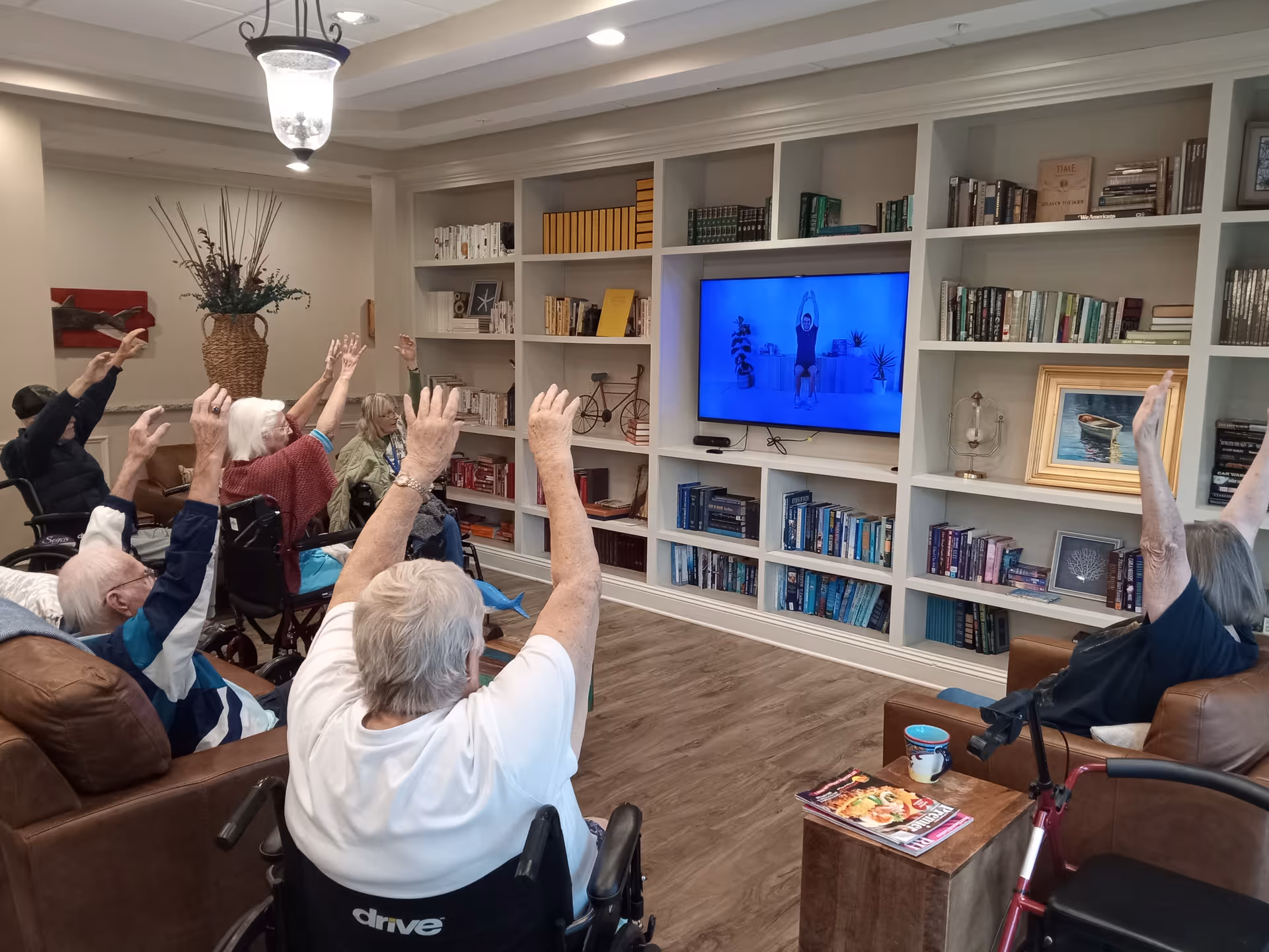 A group of elderly individuals seated in a living room area, some in wheelchairs, participating in a seated exercise session by following a person on a television screen. The room has built-in white bookshelves filled with books and decorative items, wood flooring, and comfortable seating.
