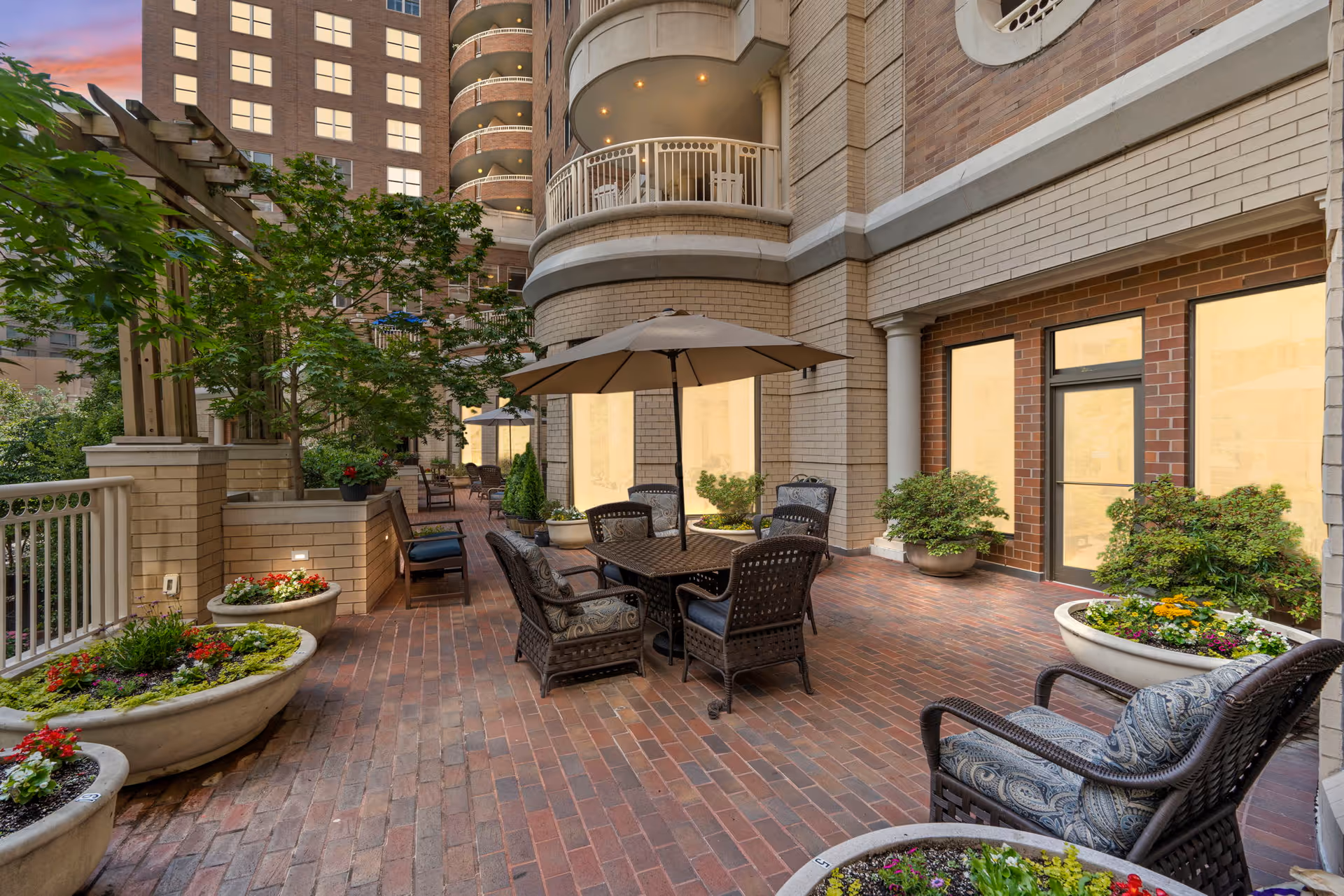 Outdoor patio area at The Jefferson featuring brick flooring, several large planters with colorful flowers and greenery, wicker chairs with cushions, tables with umbrellas, and a multi-story building with balconies in the background during sunset.