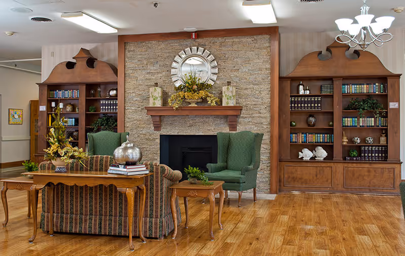 A cozy living room area in a senior living facility featuring two green armchairs and a striped sofa arranged around a wooden coffee table and side table. Behind the seating is a stone fireplace with a decorative round mirror and floral arrangements on the mantel. Flanking the fireplace are two large wooden bookshelves filled with books and decorative items. The room has hardwood flooring and a chandelier hanging from the ceiling.