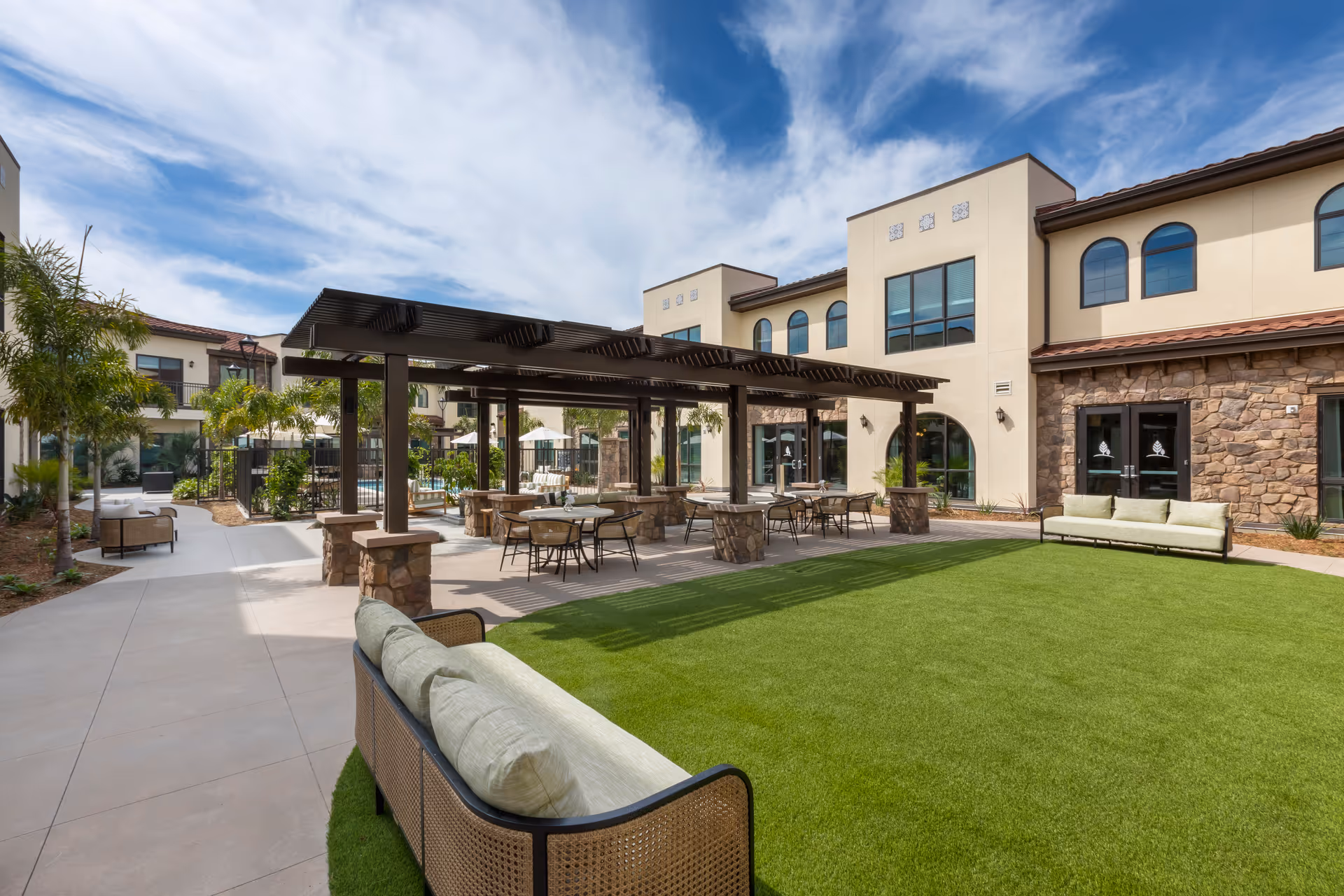 Courtyard of a senior living facility featuring a pergola with tables and chairs, lounge seating, and a green lawn in front of a two-story building.