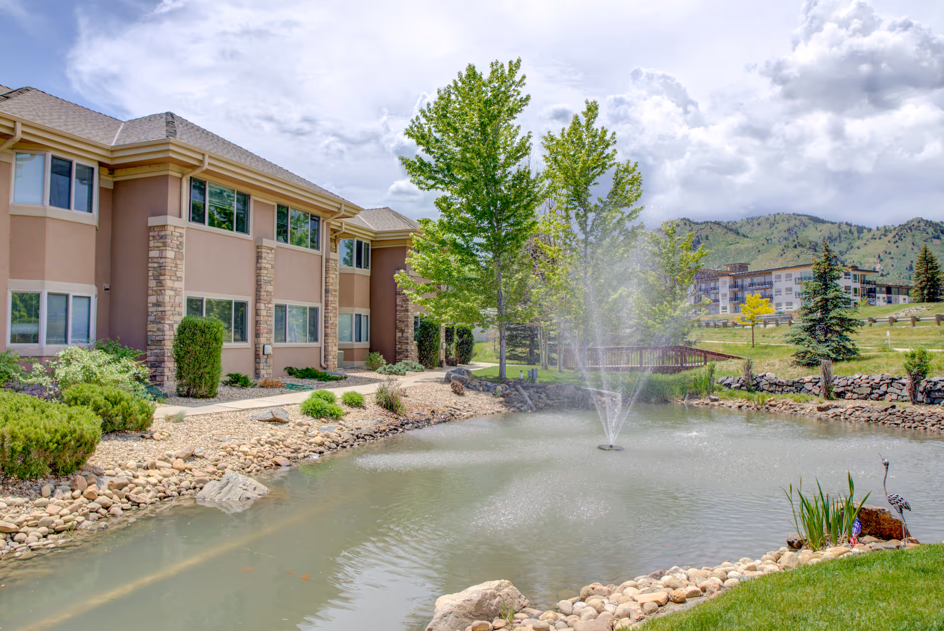 Exterior view of Golden Pond Senior Living facility showing a two-story building with beige walls and stone accents next to a pond with a water fountain. The pond is surrounded by rocks and greenery, with trees and mountains in the background under a partly cloudy sky.