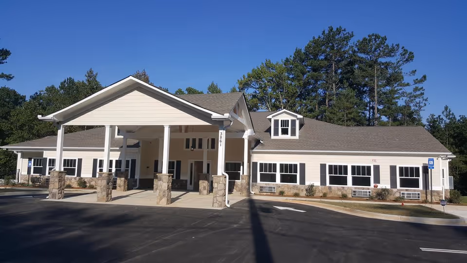 Front exterior view of a single-story senior care home building with a large covered entrance supported by white columns with stone bases. The building is light-colored with multiple windows and surrounded by trees under a clear blue sky.