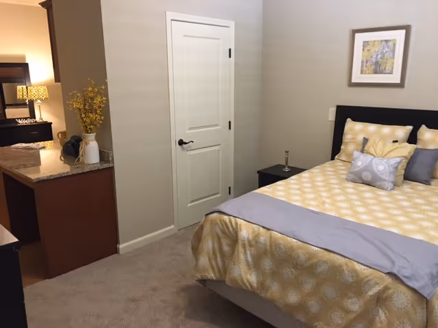 Furnished bedroom with a yellow-patterned bed, nightstand, decorative accents, and a door leading to an adjacent room.