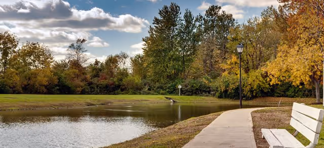 A peaceful outdoor scene featuring a small pond surrounded by green grass and trees with autumn foliage. A paved walkway curves along the right side of the pond, with a white bench and a black lamppost beside it under a partly cloudy sky.
