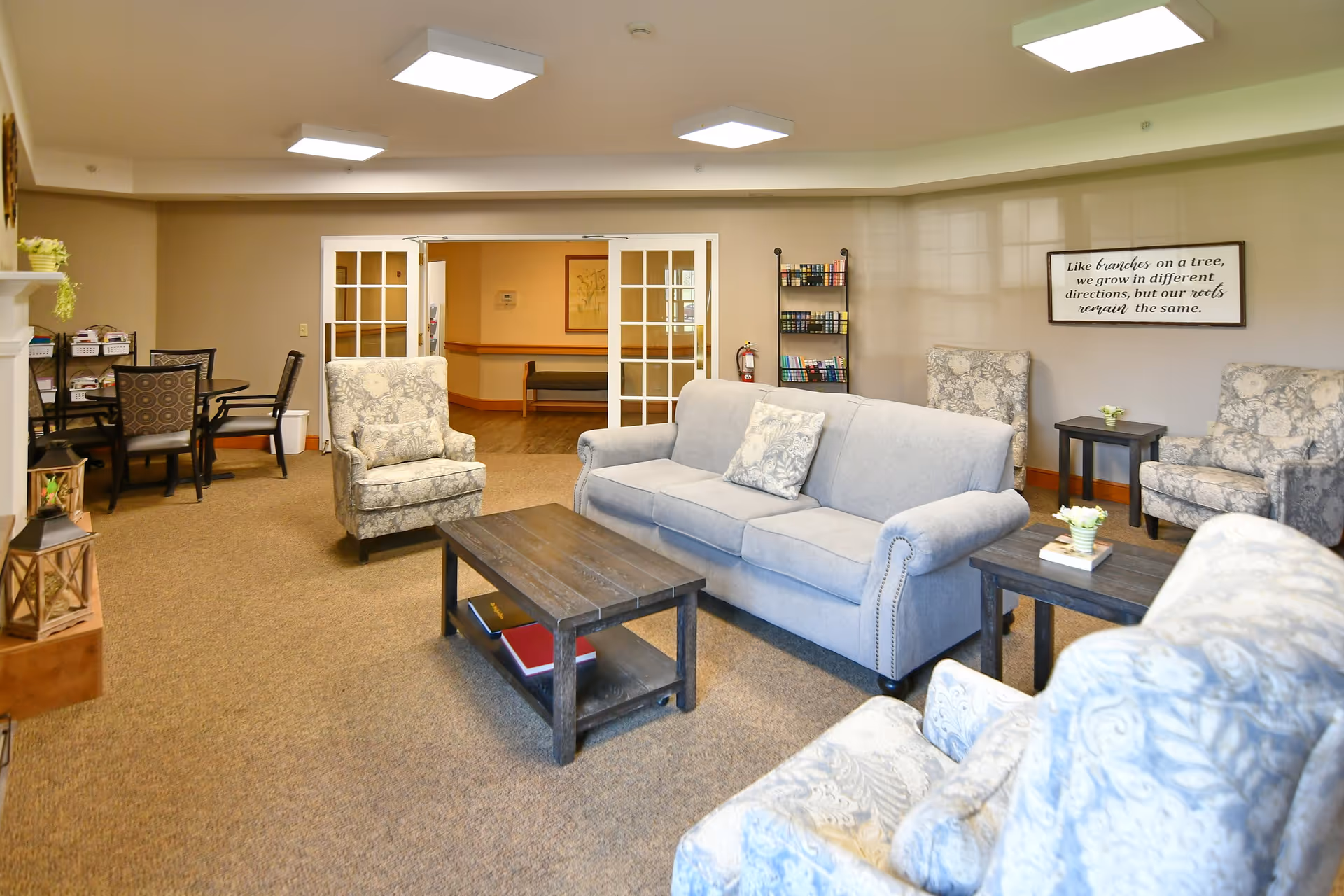 A cozy senior living facility common area with a light gray sofa, patterned armchairs, a dark wooden coffee table, and side tables with small flower arrangements. There is a bookshelf with books, a round table with chairs, and a framed inspirational quote on the wall. The room has beige walls and carpet, with bright ceiling lights and double glass doors leading to another room.