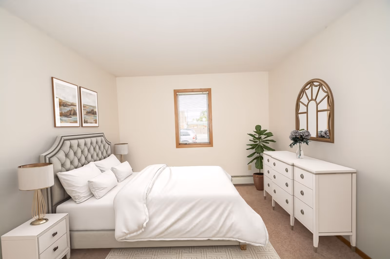 A neatly made bed with white bedding and pillows in a bedroom. The bed has a tufted gray headboard. On the left side, there is a white nightstand with a lamp. On the right side of the room, there is a white dresser with multiple drawers, a decorative mirror above it, and a vase with flowers. A potted plant is placed in the corner near the window with wooden trim. Two framed landscape paintings hang on the wall above the bed.