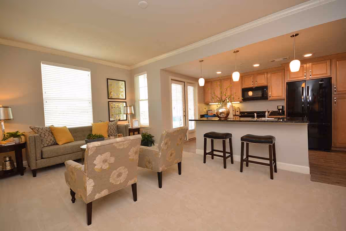 A bright and cozy living room area with a gray sofa adorned with yellow and patterned cushions, two floral-patterned armchairs, and side tables with lamps and plants. The room opens into a kitchen with wooden cabinets, black appliances, a granite countertop island with two black stools, and pendant lights hanging above.