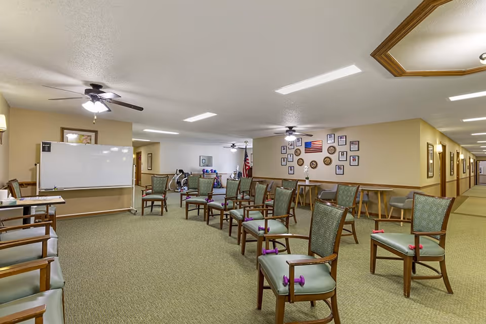 A spacious room with multiple chairs arranged in rows, each chair holding a small purple dumbbell. The room has beige walls, a green carpet, ceiling fans with lights, and a whiteboard on one wall. There are framed pictures and an American flag displayed on the far wall, with a hallway visible to the right.