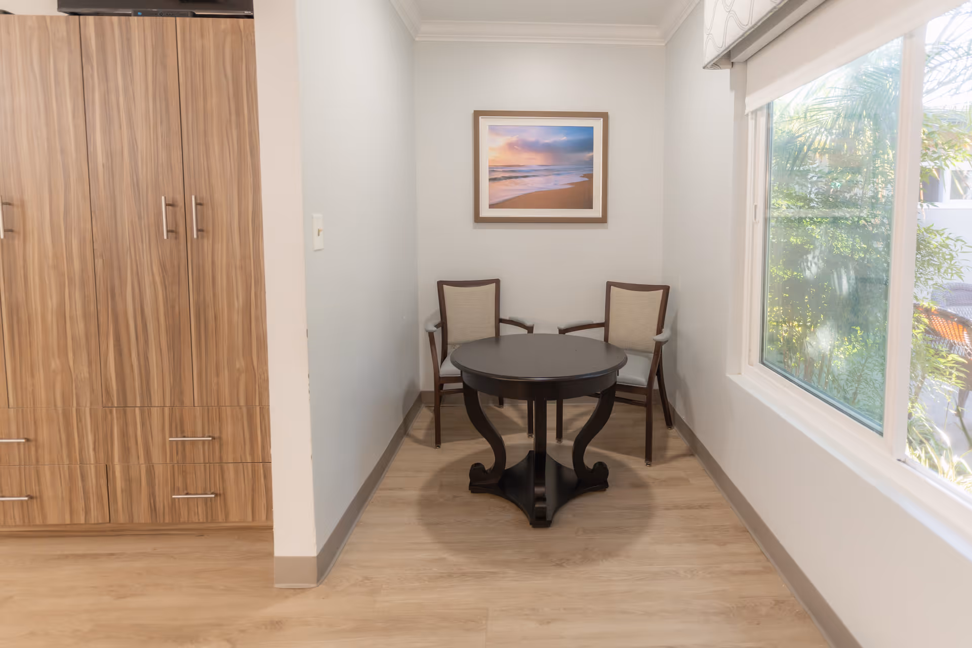 A small seating area with a round dark table and two chairs beneath a framed beach photograph, next to a large window and wood cabinets.