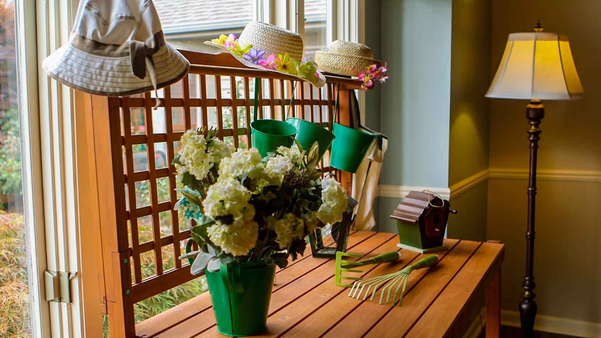 A wooden table by a window in a cozy interior holding green potted flowers, gardening tools, decorative hats, and a small birdhouse with a floor lamp nearby.