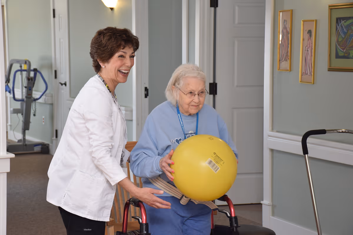 An elderly woman using a walker holds a large yellow exercise ball while a smiling female caregiver or therapist stands beside her, assisting and encouraging her in a hallway with exercise equipment visible in the background.