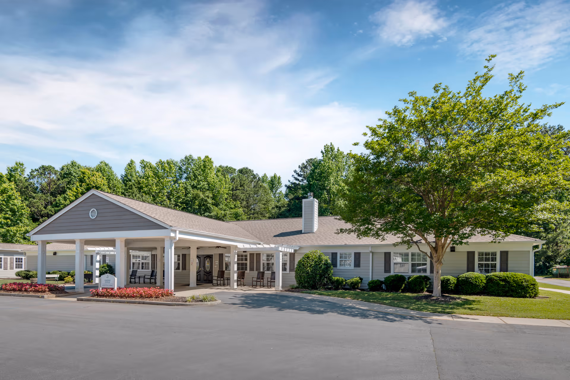 Exterior view of a single-story senior living facility building with a covered entrance, surrounded by greenery including trees and bushes under a partly cloudy blue sky.