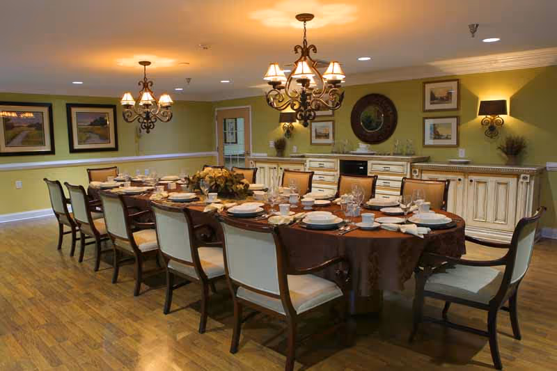 A large dining room with a long rectangular table set for a meal. The table is covered with a brown tablecloth and has place settings including plates, cups, and napkins. There are multiple chairs around the table. The room has wooden flooring, green walls, framed pictures, and two ornate chandeliers hanging from the ceiling. A sideboard with decorative items is against the far wall.