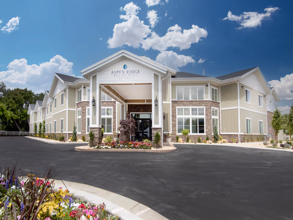 Two-story Aspen Ridge Residences of Lehi building front with a covered entrance, landscaped flower beds, and a paved driveway under a blue sky.
