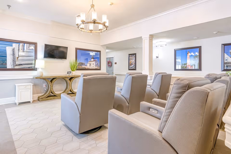 A bright and clean senior living common area with several comfortable gray recliner chairs arranged in rows facing a wall-mounted flat screen TV. The walls are decorated with framed pictures of lighthouses and coastal scenes. A wooden console table with a lamp and a plant is positioned beneath the TV. The room has light-colored tile flooring and a modern chandelier hanging from the ceiling.