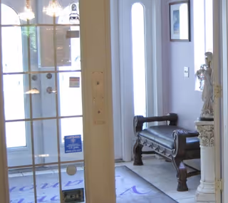 Interior view of an entrance area with a glass door, a decorative bench with dark wood and black cushion, a statue on a pedestal, and framed artwork on the wall. Light is coming through the glass door and windows.