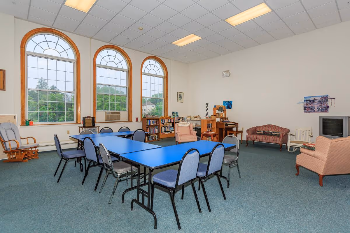 A spacious common room with three large arched windows letting in natural light. The room features blue tables arranged in a U-shape with several chairs around them. There are bookshelves filled with books, a rocking chair, a striped sofa, armchairs, a TV on a stand, and various decorations on the walls. The carpet is green, and the ceiling has recessed lighting.
