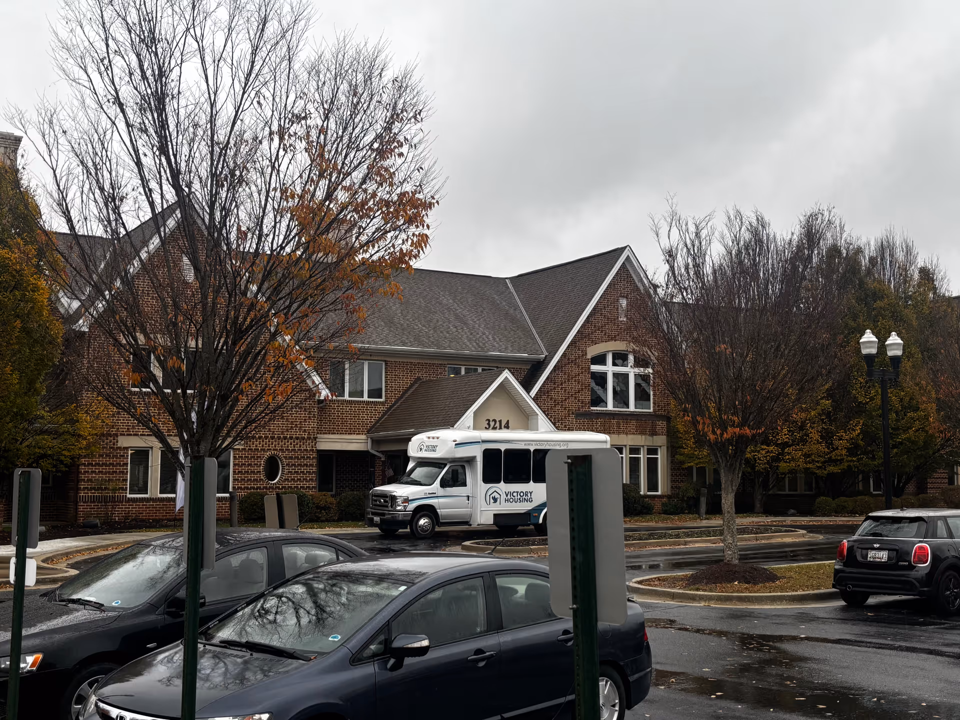Exterior view of a brick senior living facility named Grace House on a cloudy day with several parked cars and a white shuttle bus labeled Victory Housing in front of the building. Trees with sparse autumn leaves are visible around the parking lot.