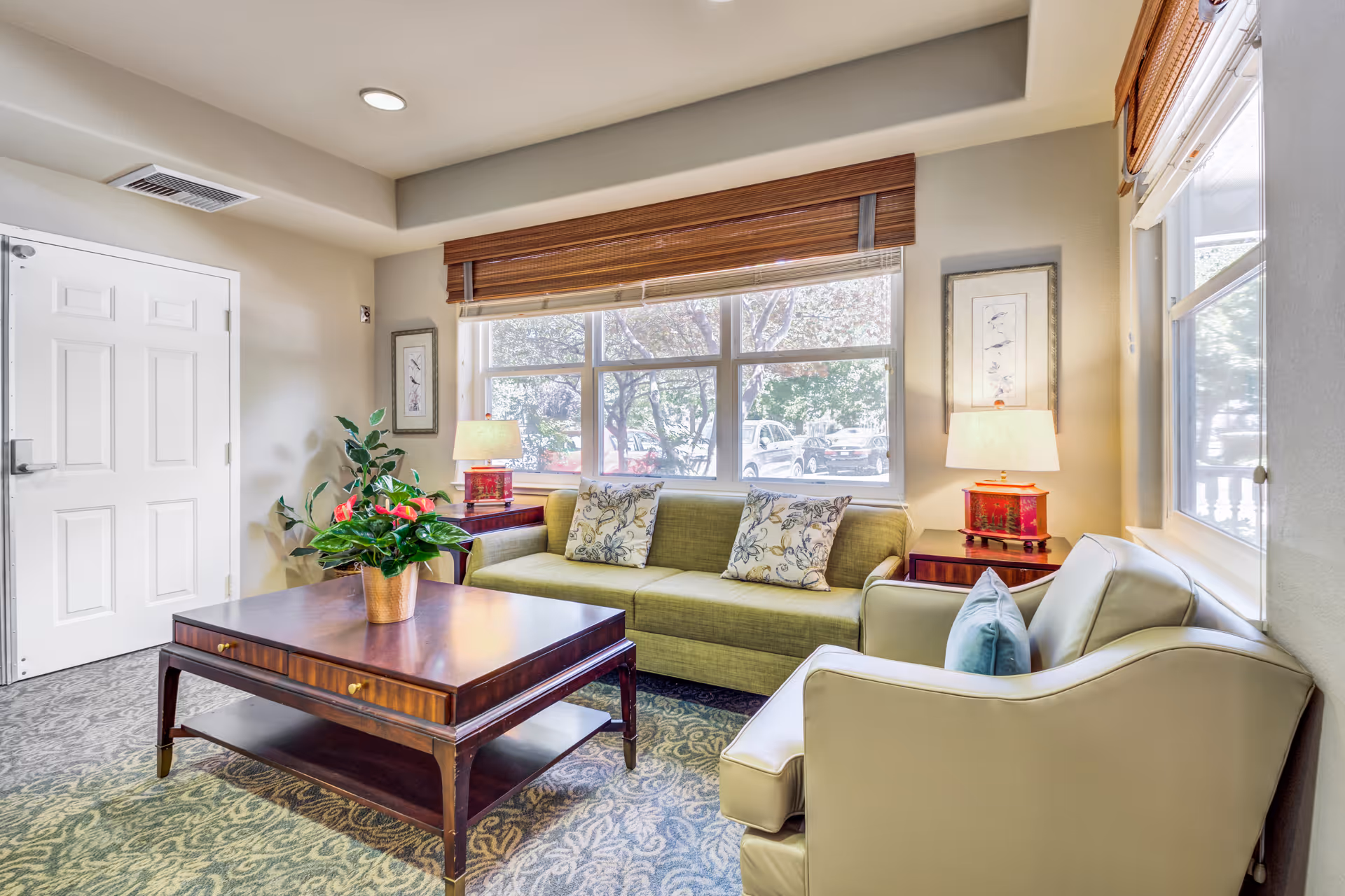 A bright and cozy living room with a green sofa adorned with patterned cushions, a beige armchair with a blue pillow, a wooden coffee table with a potted plant, two side tables with lamps, and large windows letting in natural light.