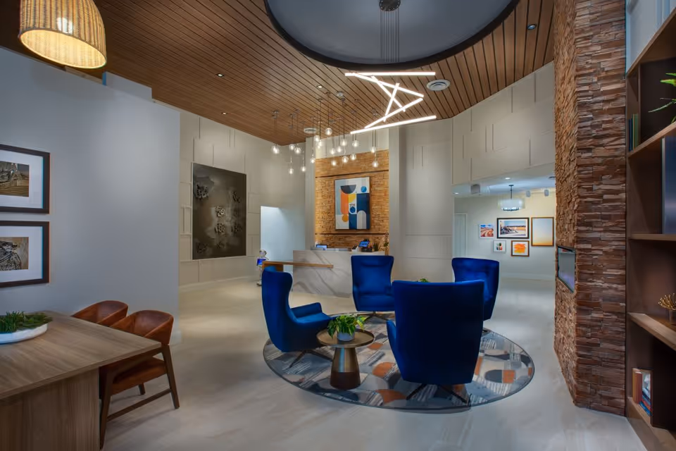 A modern and stylish senior living facility lobby with a circular seating area featuring four blue armchairs around a small round table on a patterned rug. The space has a wooden ceiling with contemporary light fixtures, a reception desk with a colorful abstract painting behind it, and walls decorated with framed artwork. There is a wooden table with two brown chairs on the left and a stone accent wall with built-in shelving on the right.