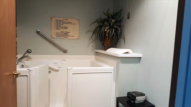 A small bathroom with a walk-in bathtub featuring a door for easy access. There is a metal grab bar on the wall above the tub, a faucet on the left side, and a sign with instructions mounted on the wall. A potted plant is placed on a ledge next to the tub, and a small black table with a device on it is visible in the corner.