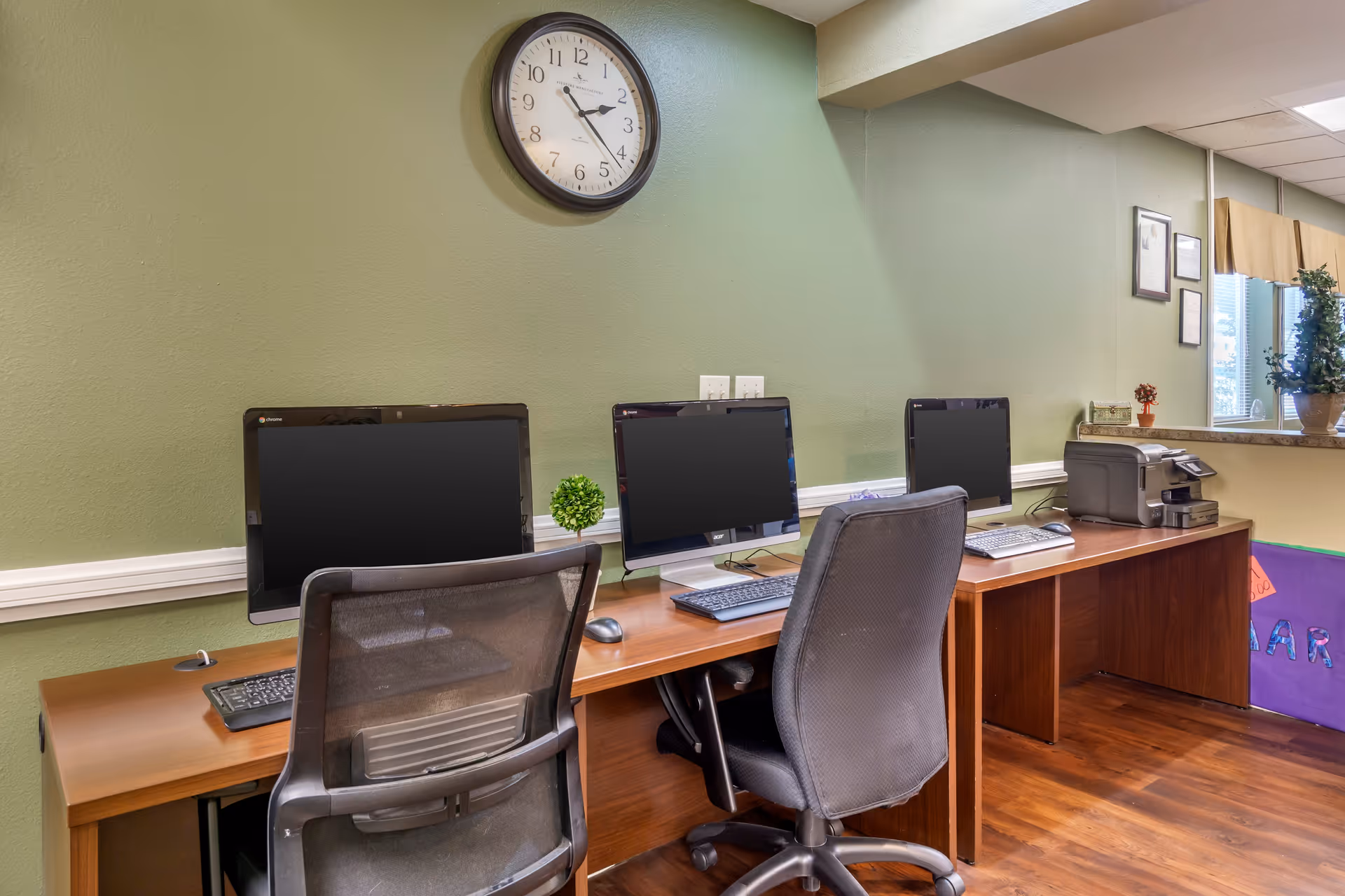 A computer workstation area with three desktop computers on a wooden desk against a green wall. Two office chairs are positioned in front of the computers. A round wall clock above the desk shows the time as 2:25. There is a printer on the right side of the desk and a small decorative plant between two of the monitors. The room has wooden flooring and a window with a potted plant on the windowsill.