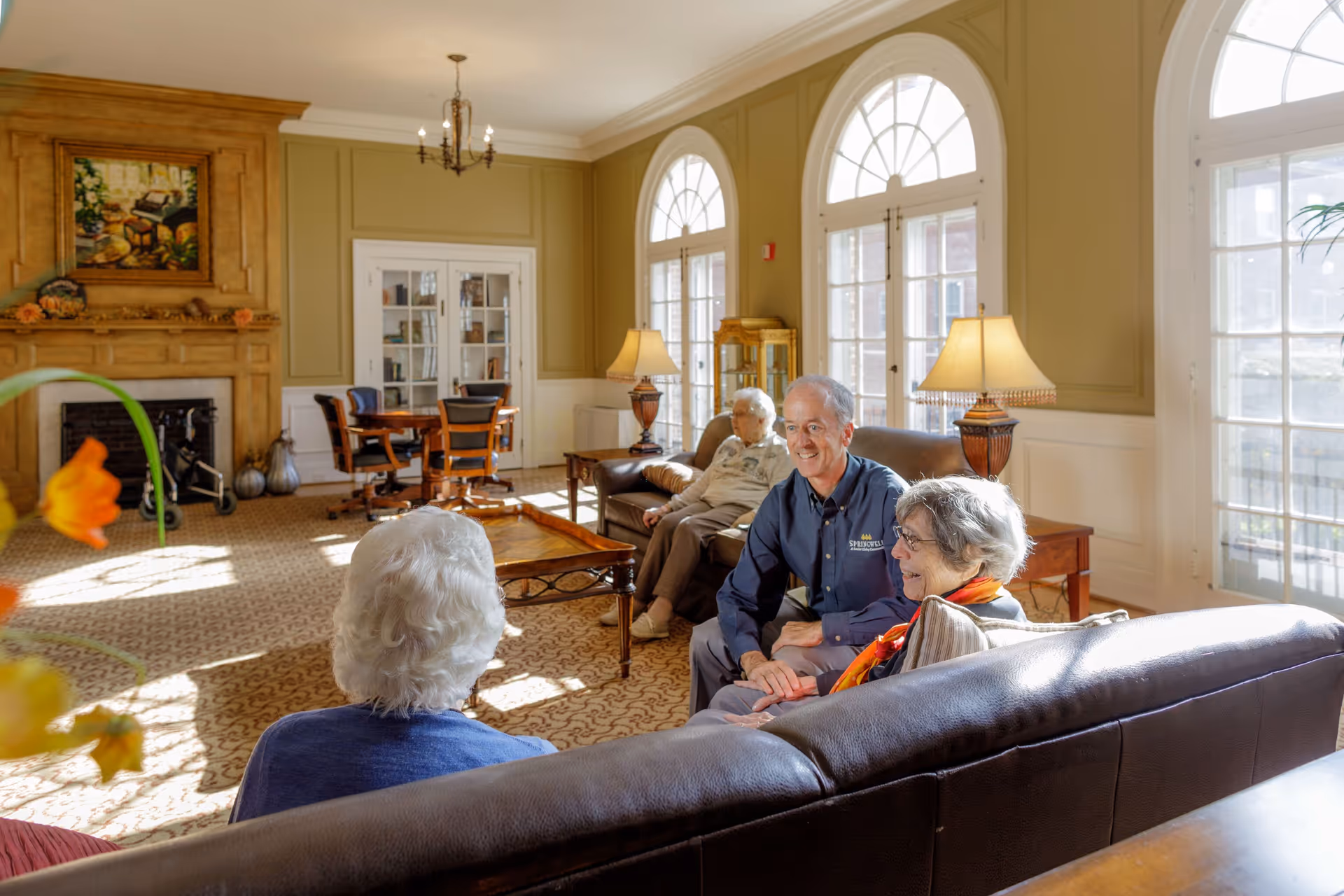 A bright and spacious living room in a senior living community with large arched windows allowing natural light to fill the room. There are three elderly people and one staff member sitting and conversing on leather sofas. The room features a fireplace with a painting above it, a patterned carpet, wooden furniture, and table lamps providing a warm ambiance.