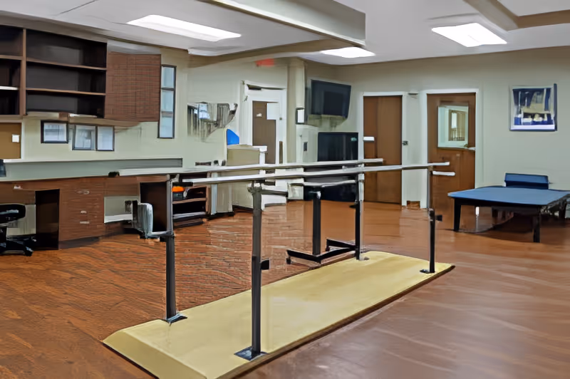 A rehabilitation room with parallel walking bars on a padded platform, desks, and a treatment table.