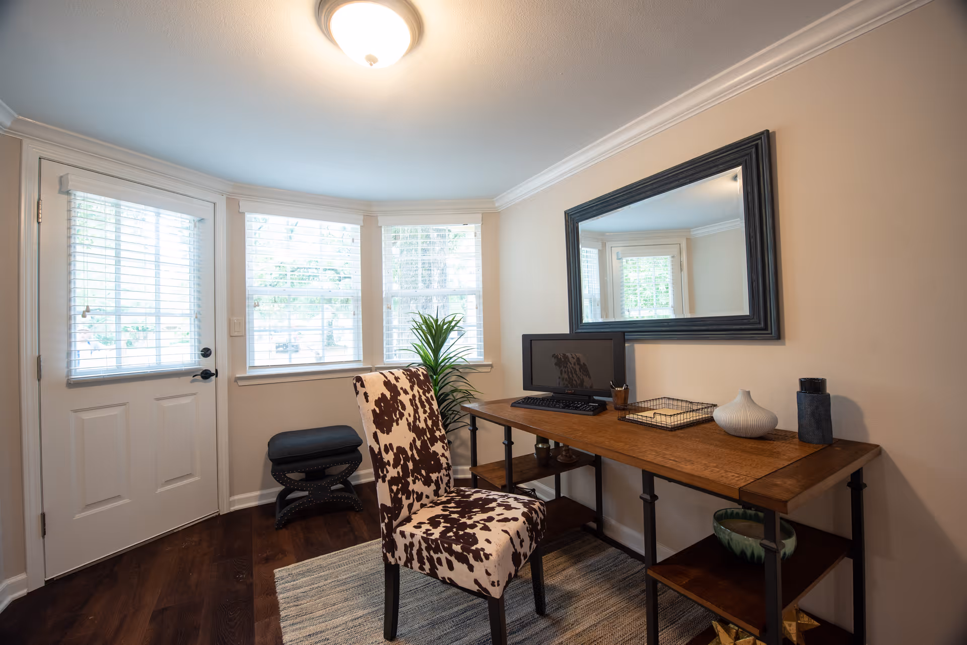 A small home office area with a wooden desk holding a computer monitor, keyboard, pencil holder, decorative items, and a wire tray. A chair with a brown and white cowhide pattern is placed in front of the desk. The room has three windows with white blinds and a door with a window, letting in natural light. A large rectangular mirror hangs on the wall above the desk, and a small plant is positioned near the windows. The floor is dark wood with a light-colored rug under the desk and chair.