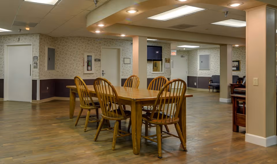 Interior view of a common area in a nursing center featuring a wooden table with six wooden chairs on a wood floor. The walls have a two-tone design with floral wallpaper on the upper half and dark paneling on the lower half. There are several doors, a fire extinguisher, a wall-mounted TV, and some seating visible in the background under fluorescent ceiling lights.