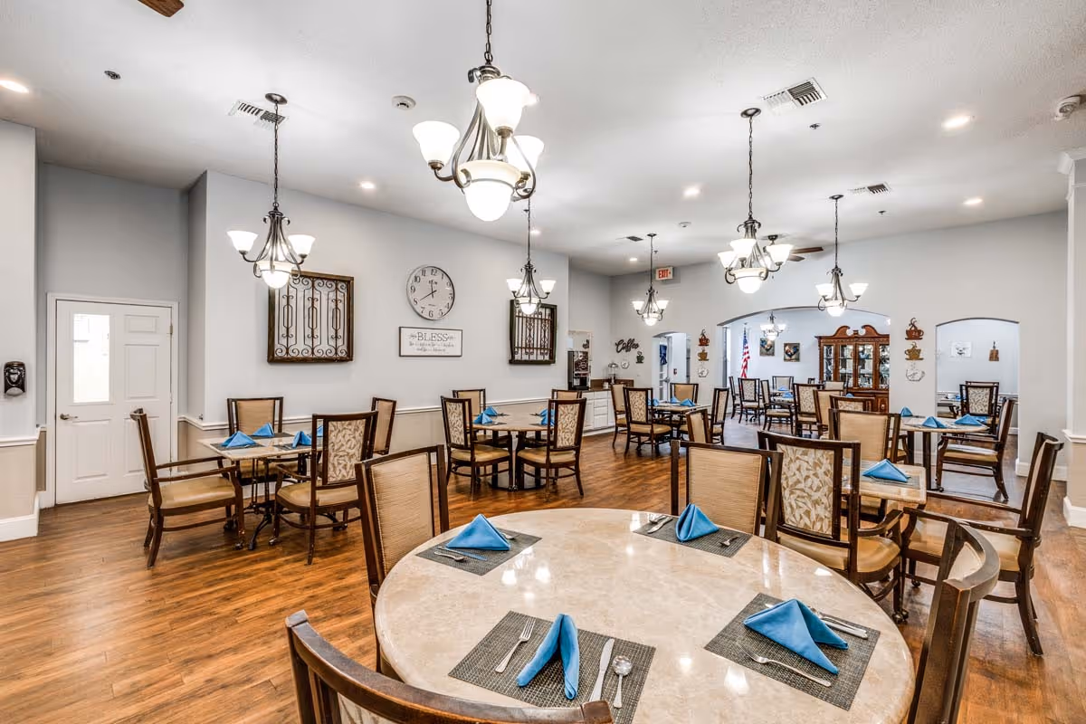 A spacious dining room in Cambridge Court Senior Living with multiple round and rectangular tables set with blue folded napkins, placemats, and silverware. The room has wooden flooring, beige walls, decorative wall art, and several hanging light fixtures providing warm lighting. There is a clock and a sign on the wall, and an exit door is visible in the background.