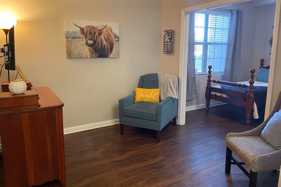 A cozy interior space featuring a wooden dresser with a lamp and decorative items, a blue armchair with a yellow pillow that says 'Grateful', and a view into a bedroom with a wooden bed frame and a window with curtains. The floor is dark wood, and the walls are light-colored with a picture of a highland cow hanging above the armchair.