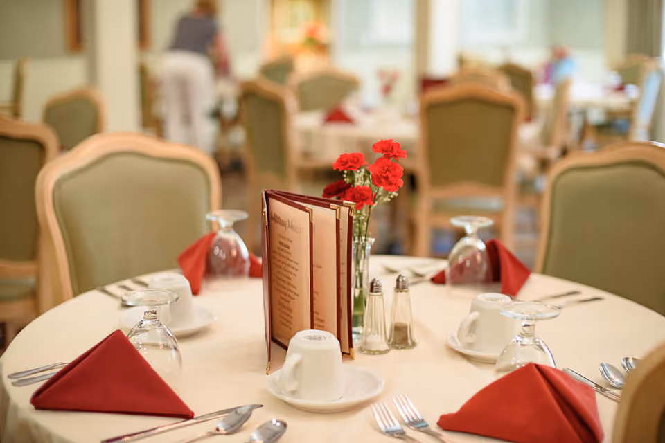 Round dining table set with menus, red napkins, overturned glasses, coffee cups and a small vase of red flowers in a softly lit dining room.