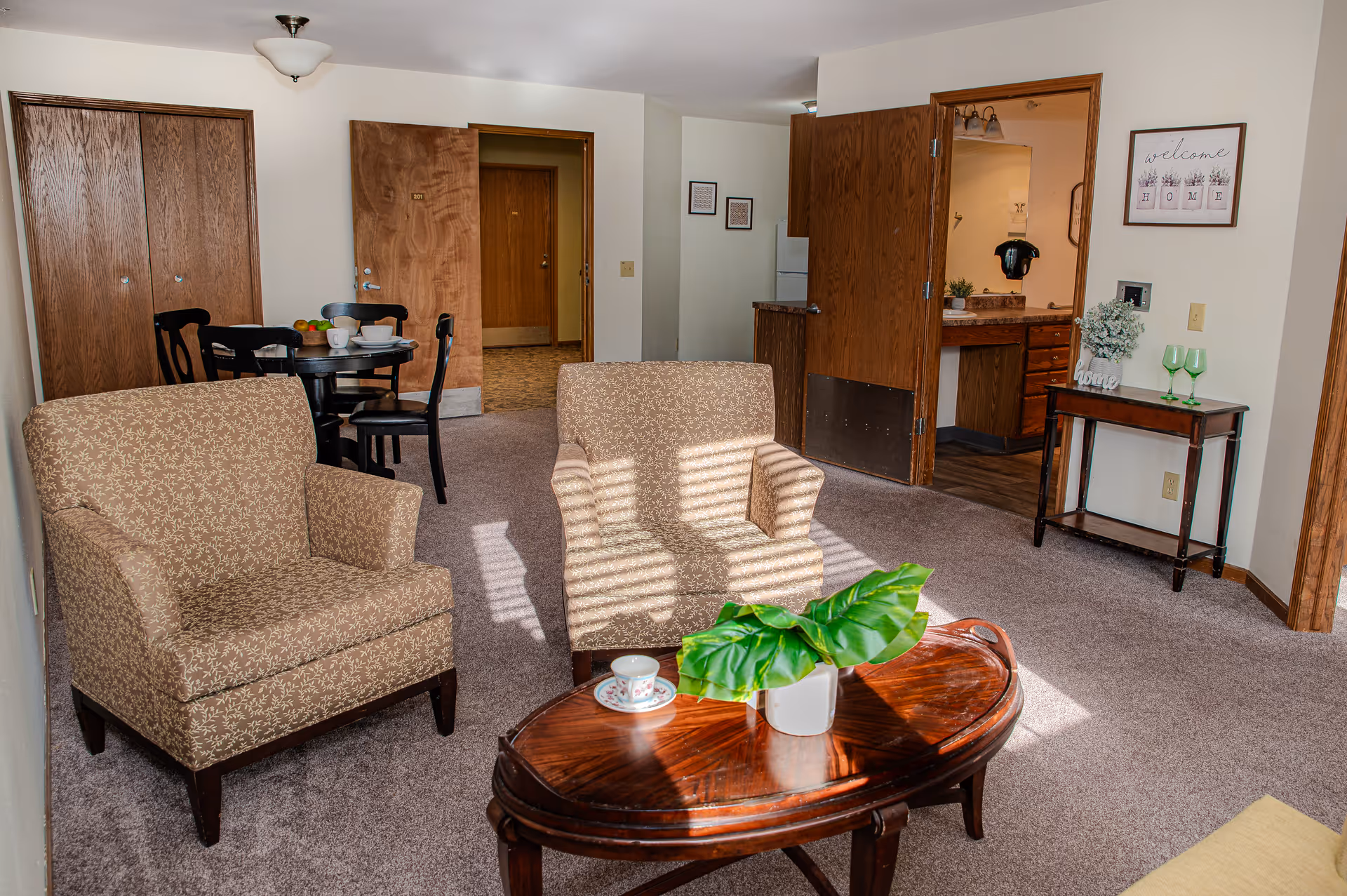 Well-lit living area with two patterned armchairs, a wooden coffee table with a potted plant, and a dining table and kitchen visible in the background.