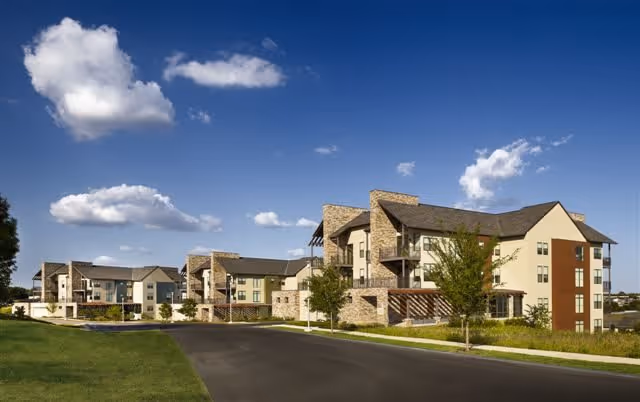 Exterior view of a modern senior living facility with multiple three-story buildings featuring stone and beige siding, surrounded by green lawns and trees under a blue sky with scattered clouds.