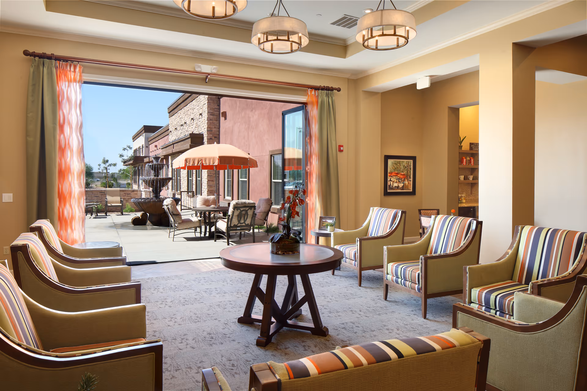 A bright and spacious living room area with multiple striped armchairs arranged around a round wooden table. Large sliding glass doors open to an outdoor patio with tables, chairs, and an umbrella. The room features warm beige walls, patterned curtains, and modern ceiling lights.
