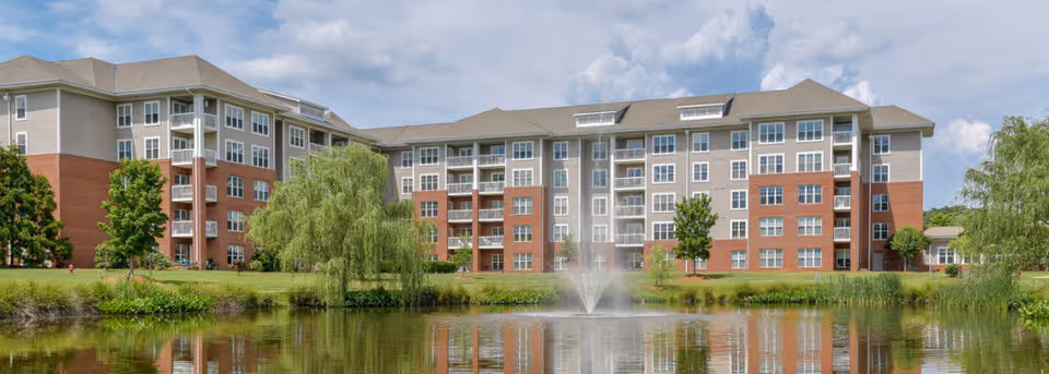 A large multi-story senior living facility building with a combination of brick and light-colored siding exterior. The building is situated behind a pond with a water fountain in the center, surrounded by green grass, trees, and shrubs under a partly cloudy sky.