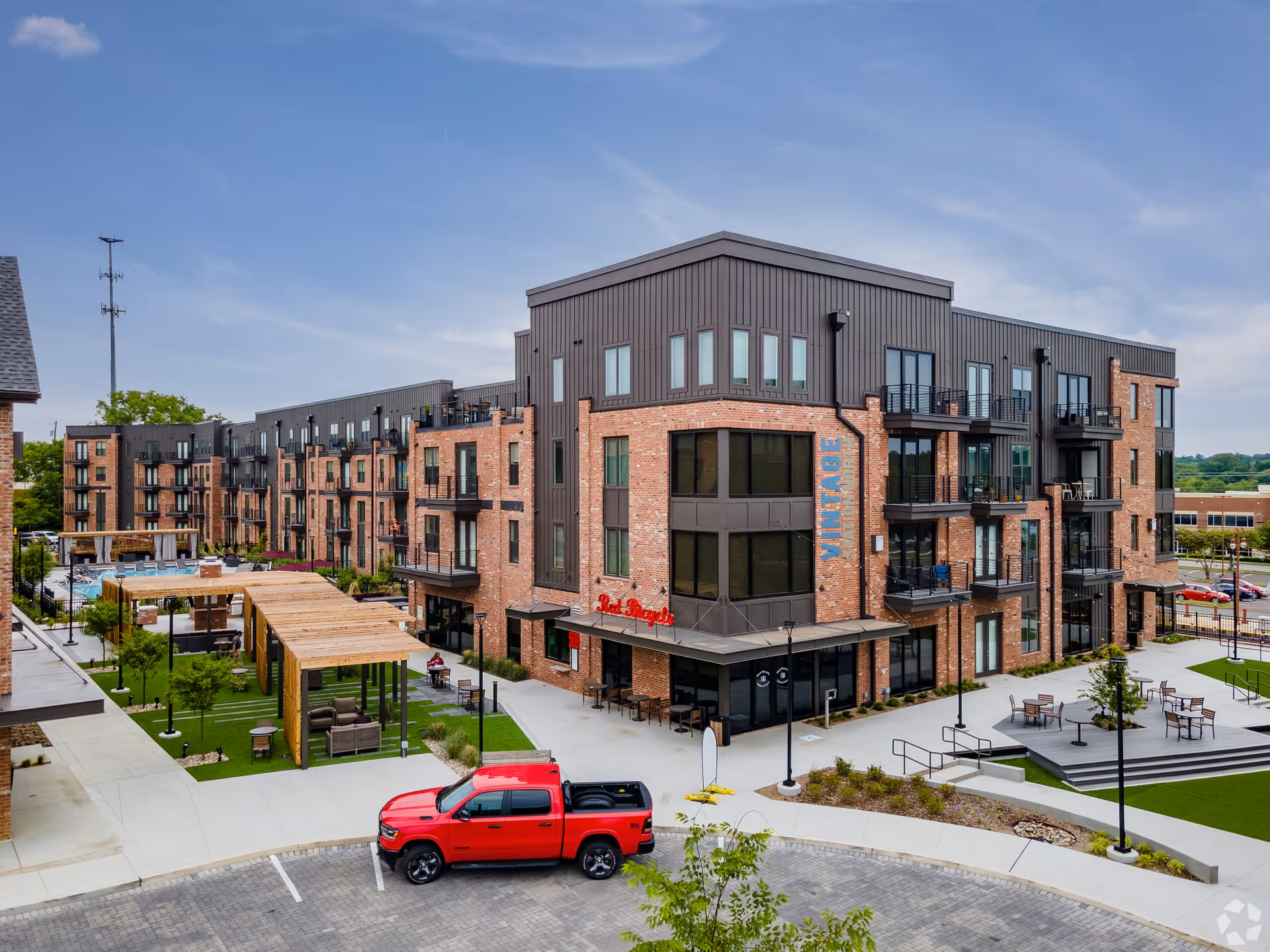 Exterior view of a modern multi-story brick and metal building with balconies, outdoor seating areas, a pergola with furniture, and a red pickup truck parked in front under a clear blue sky.